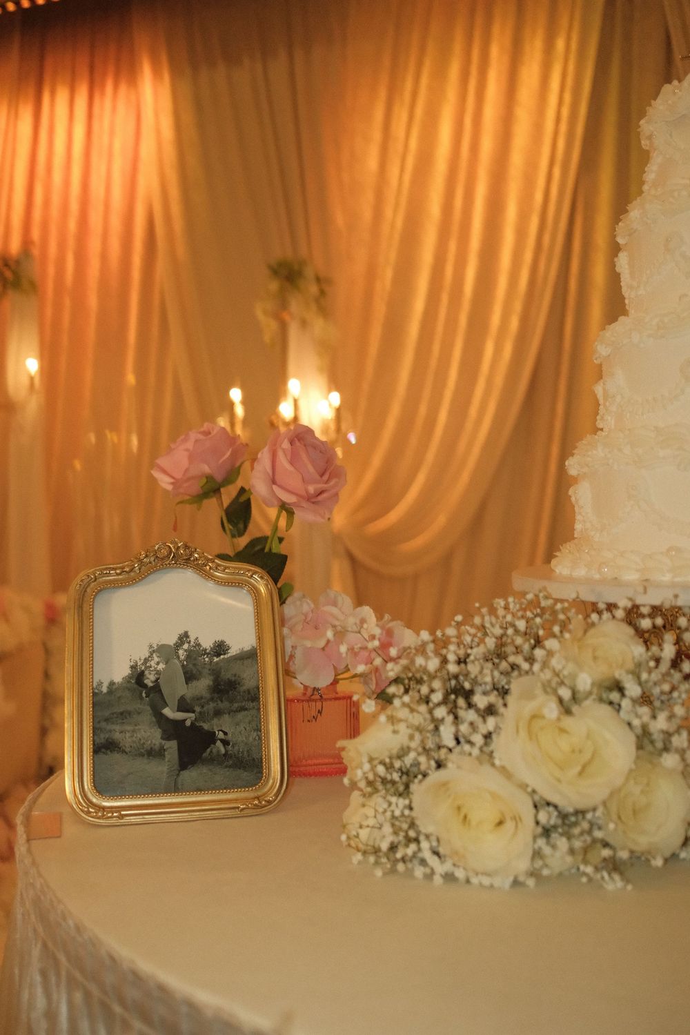 Elegant wedding cake display with white roses, baby's breath, framed black-and-white photo, pink roses, and golden candlelit drape backdrop.