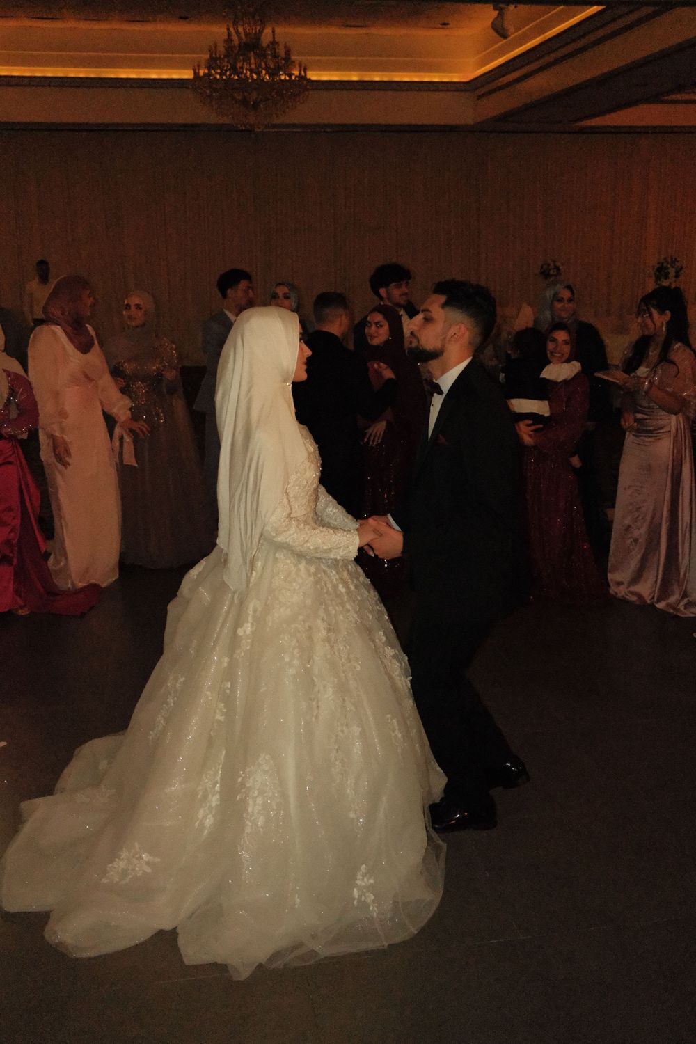 A bride in a white lace gown and veil shares a first dance with her groom in a black suit at a wedding reception.