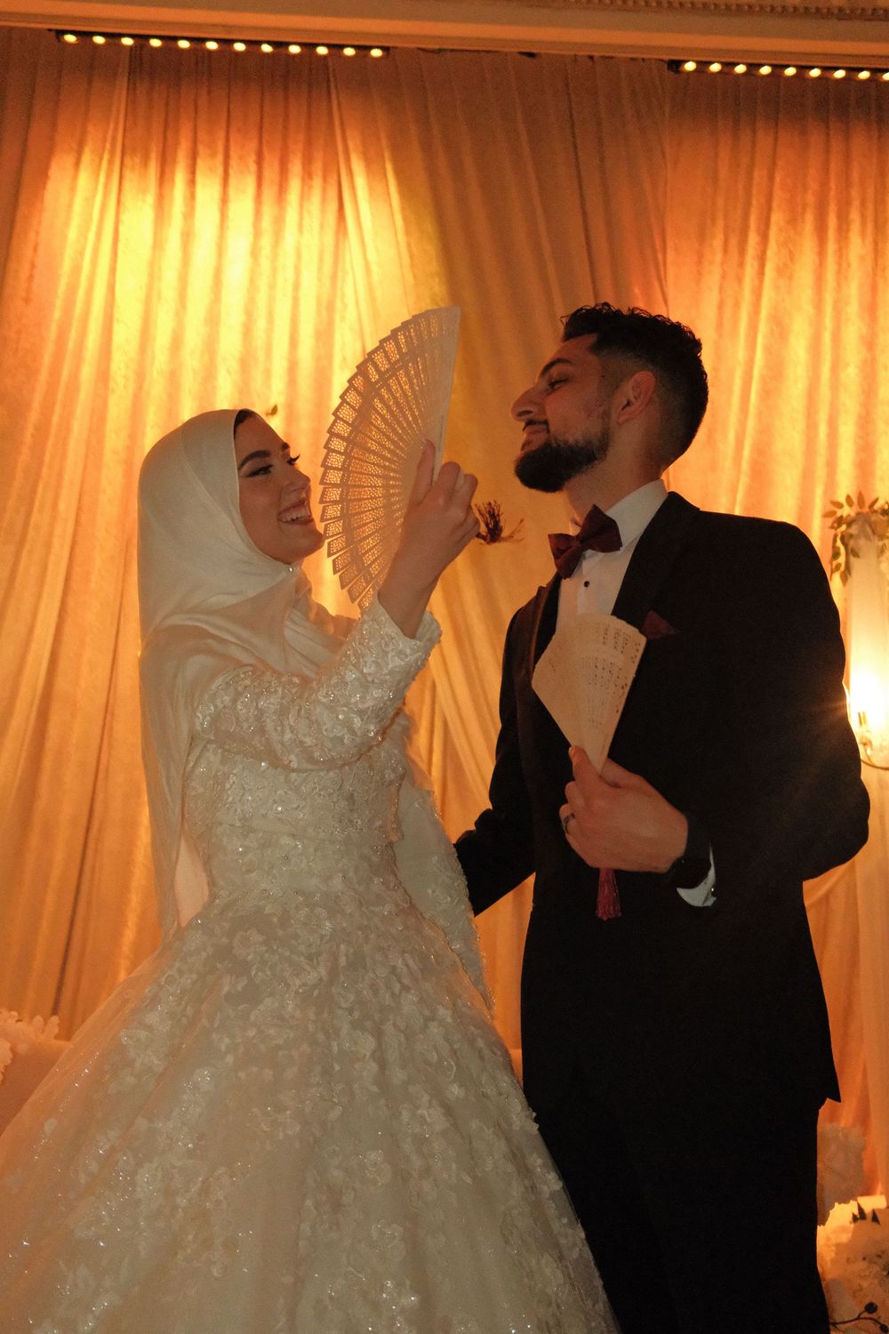 Muslim bride in white hijab and gown holds fan while groom in black tuxedo stands beside her at wedding.