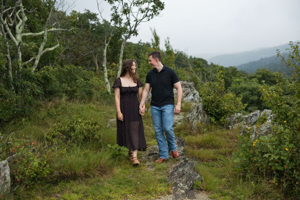 Couple holding hands on a rocky overlook surrounded by lush greenery and misty mountain views.