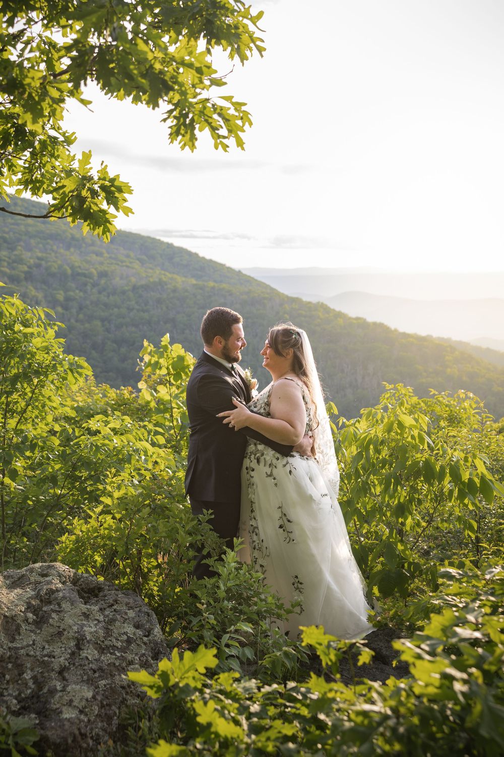 A bride and groom embrace on a mountain overlook surrounded by lush greenery at golden hour.