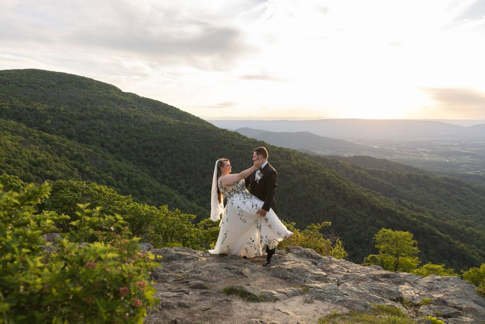 Bride and groom dancing on a rocky mountain overlook at sunset with lush green hills in the background.