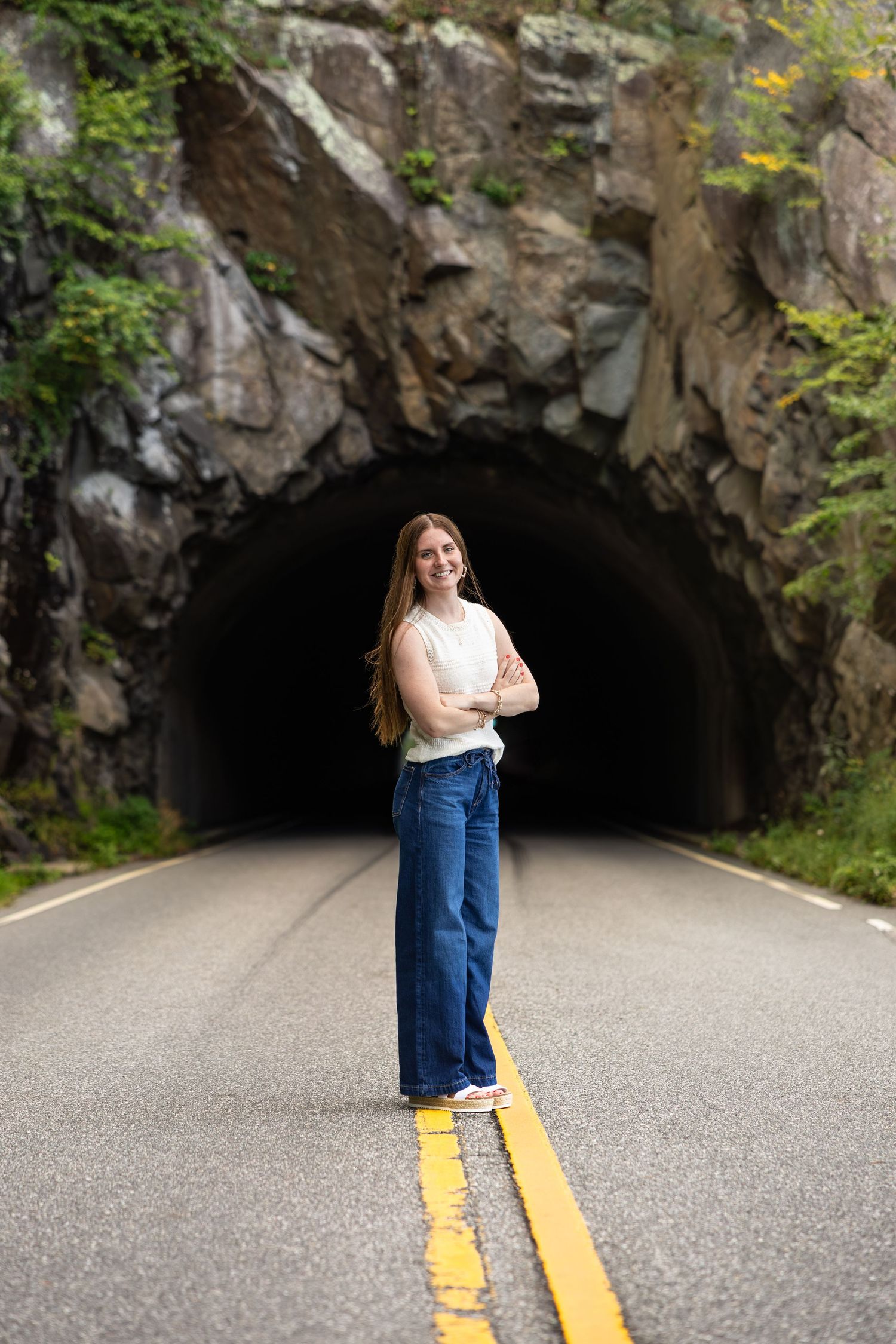 High school senior girl poses in white top and wide-leg jeans on the road in front of Mary's Rock Tunnel at the overlook in Shenandoah.