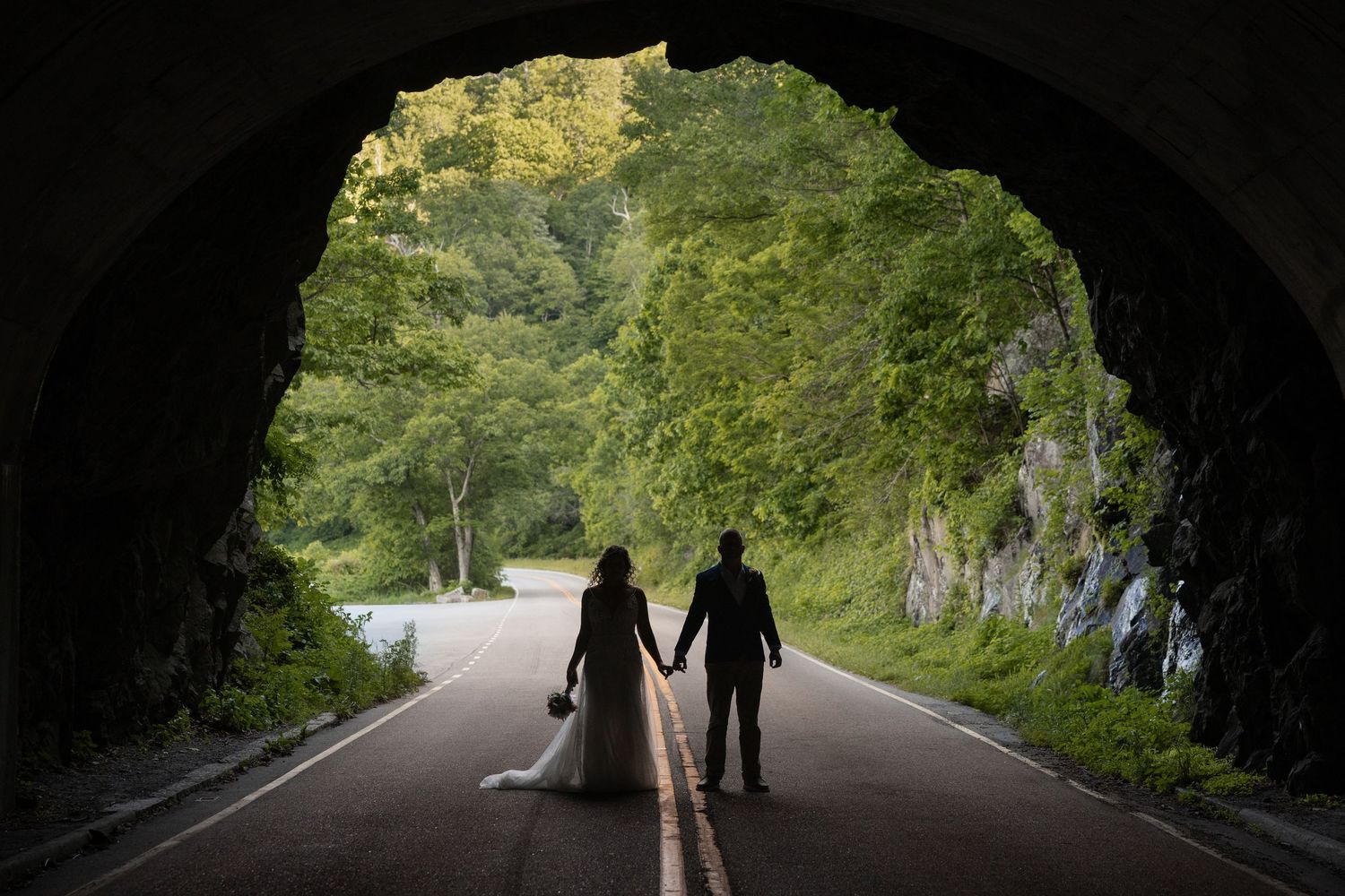 Bride and groom holding hands silhouetted inside Mary's Rock Tunnel opening with lush green trees and a winding road beyond.
