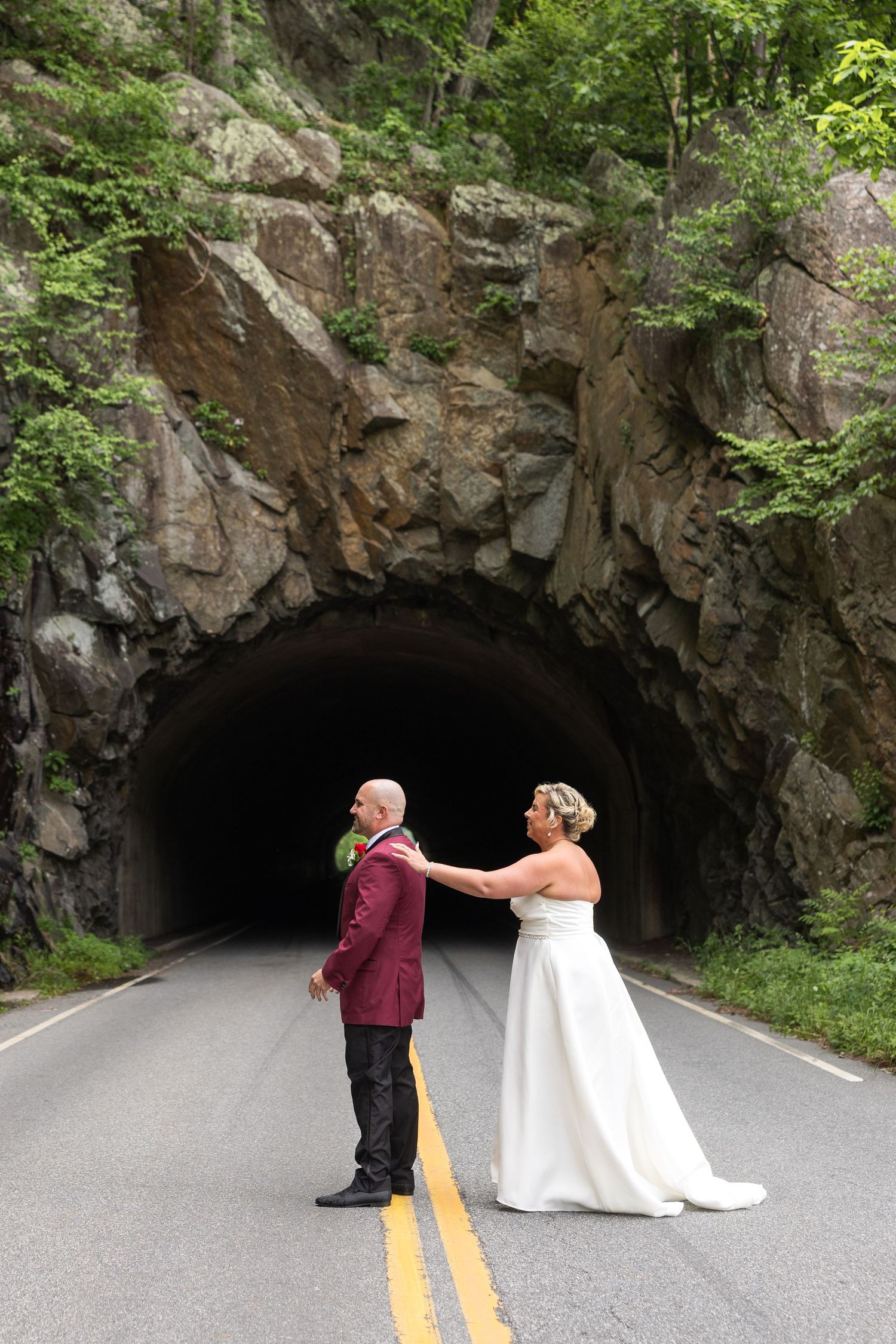 Bride and groom stand on a road before a rock tunnel entrance, surrounded by lush green trees, in a romantic wedding portrait.