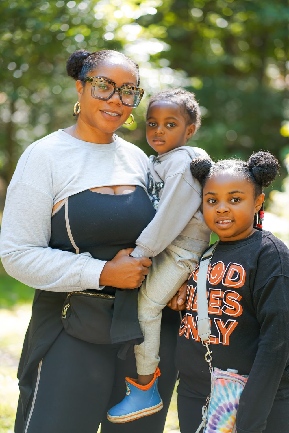 A smiling mother wearing glasses holds a toddler while standing beside an older child in a park.
