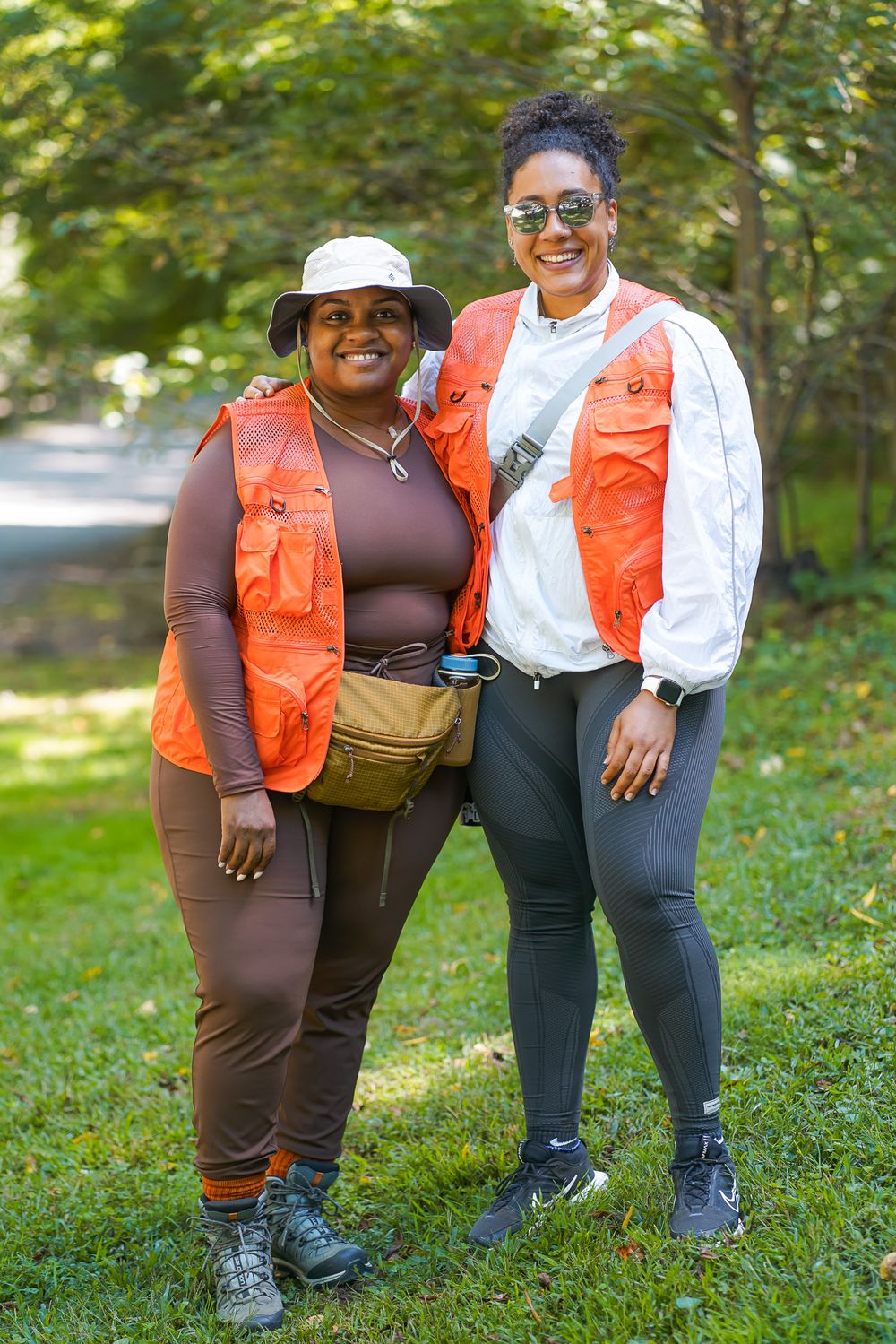 Two women wearing matching orange safety vests smile together in a green outdoor park setting.