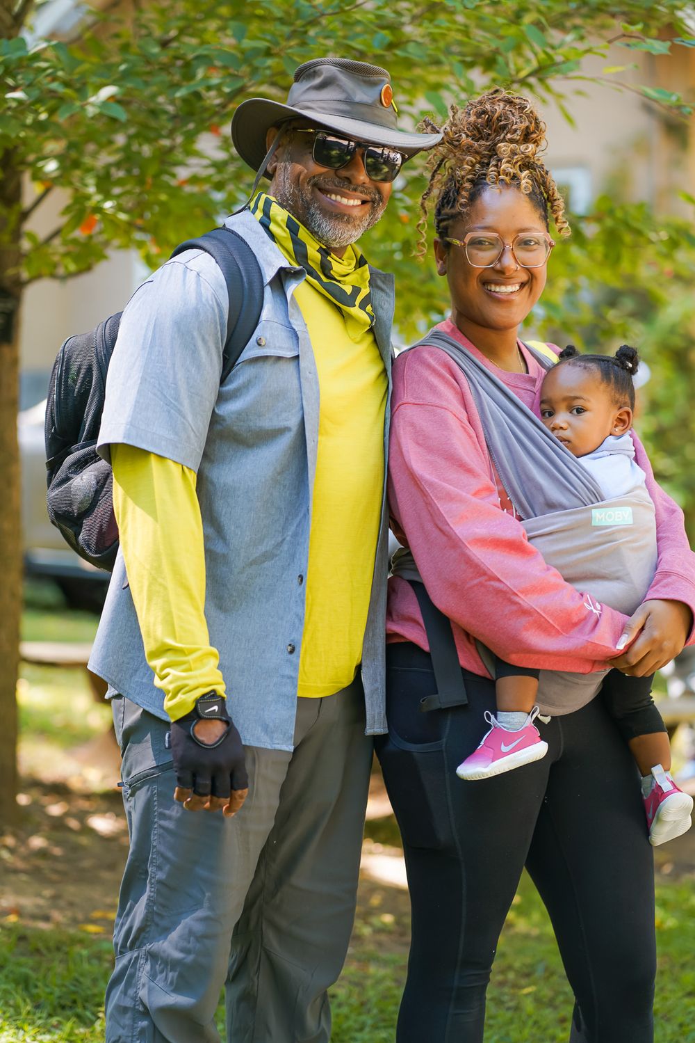 A smiling family of three poses outdoors, with a man in hiking gear and a woman carrying a baby in a carrier.