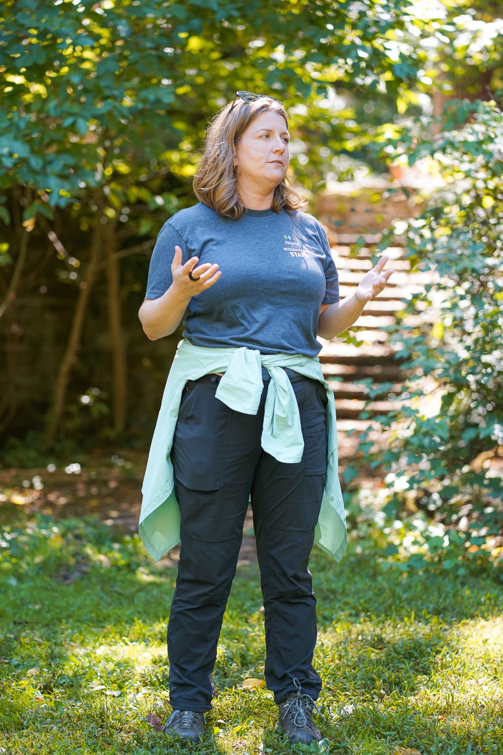 Woman in gray t-shirt gestures while speaking outdoors in a lush green garden setting.