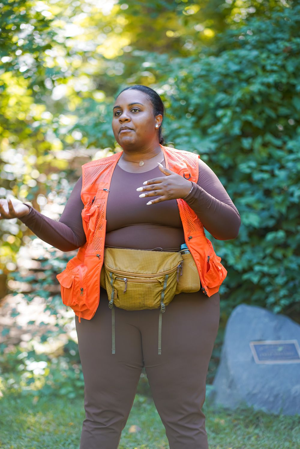 A plus-size Black woman in an orange vest, brown top, and yellow fanny pack stands outdoors gesturing.