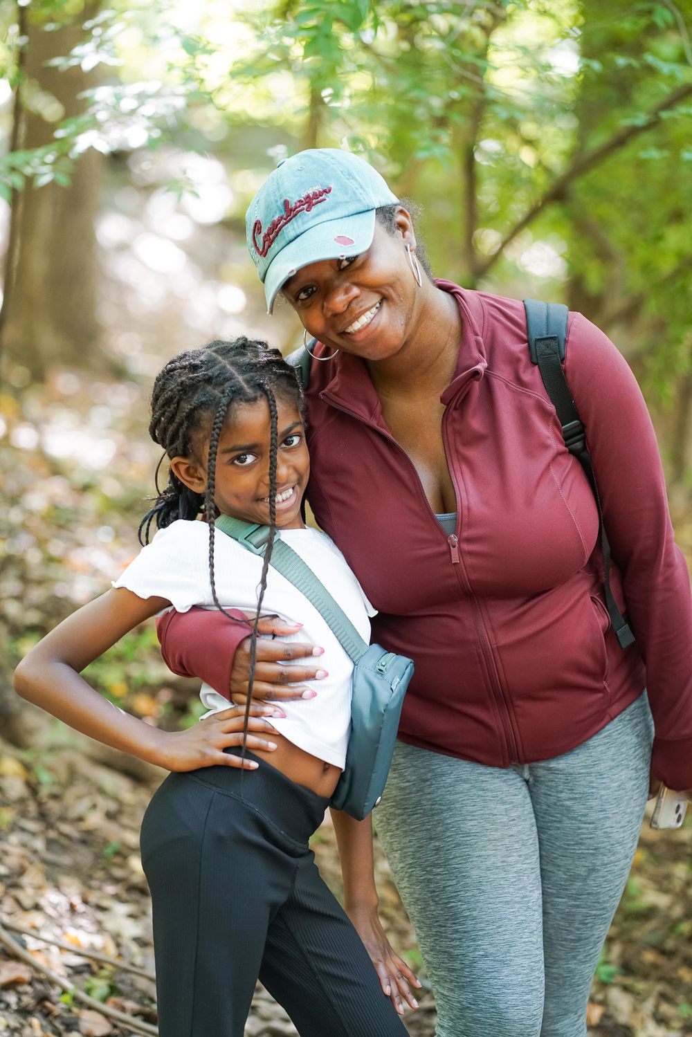 A woman and young girl smile together on a forest hiking trail, both wearing casual outdoor clothing.