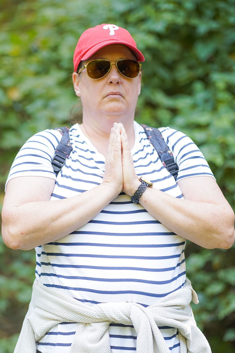 Woman in striped shirt, red cap, and sunglasses standing outdoors with hands pressed together in prayer pose.