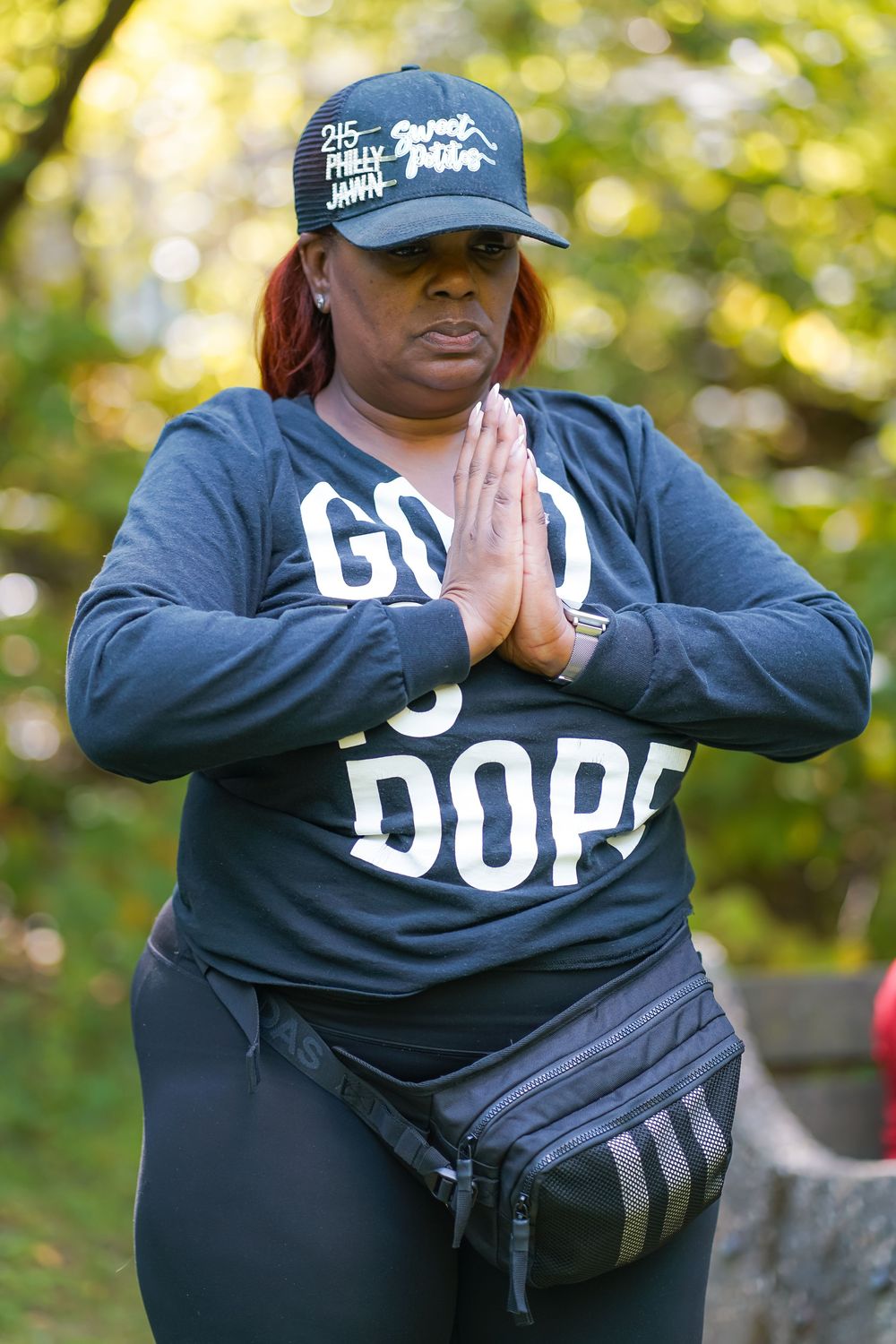 Woman in navy Good is Dope shirt and cap standing outdoors with hands pressed together in meditation pose.