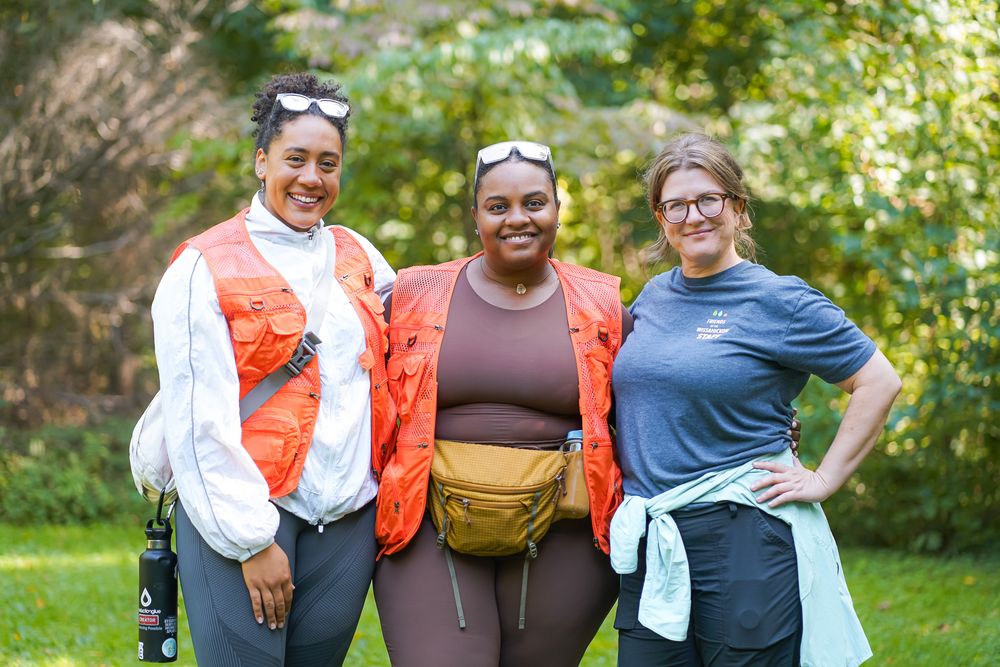 Three women smiling outdoors in hiking gear, wearing vests and fanny packs, posing together in a green park setting.