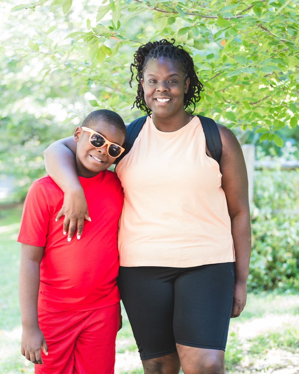 Smiling mother and son pose together outdoors, boy wearing red outfit and orange sunglasses.