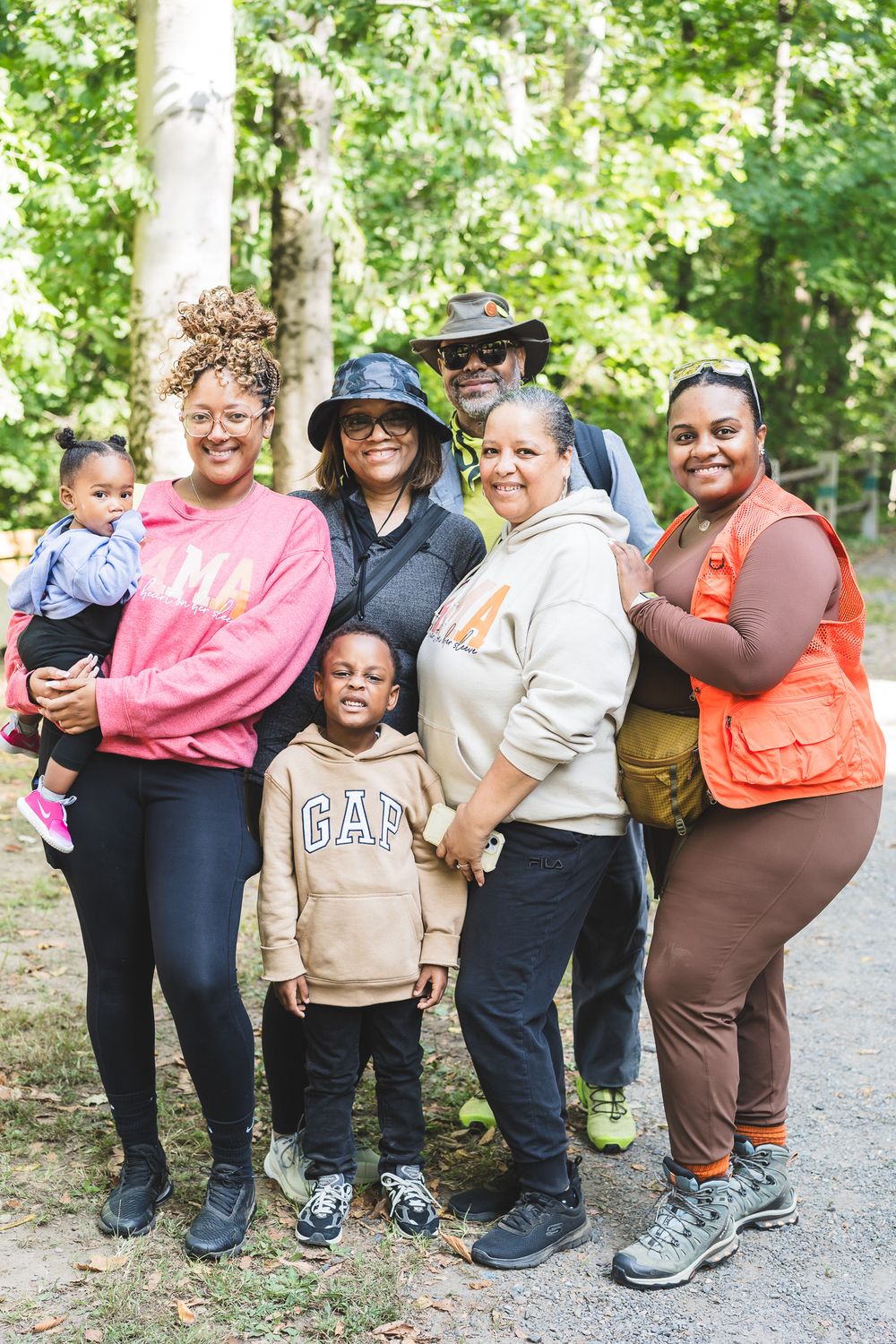 Multi-generational Black family smiling together outdoors on a nature trail surrounded by green trees.
