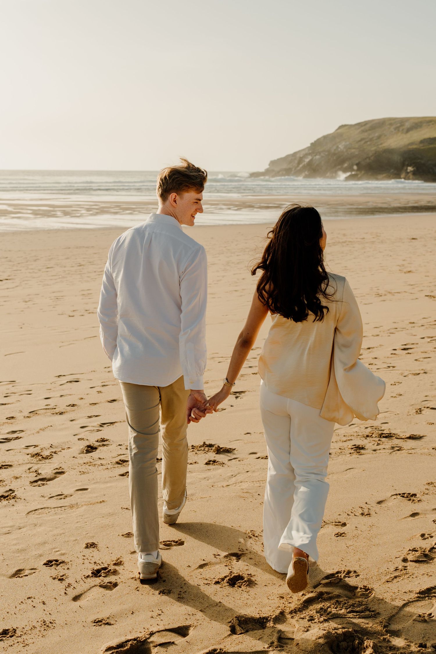Couple holding hands walking on a sandy beach at sunset, man in white shirt and woman in cream outfit with ocean backdrop.