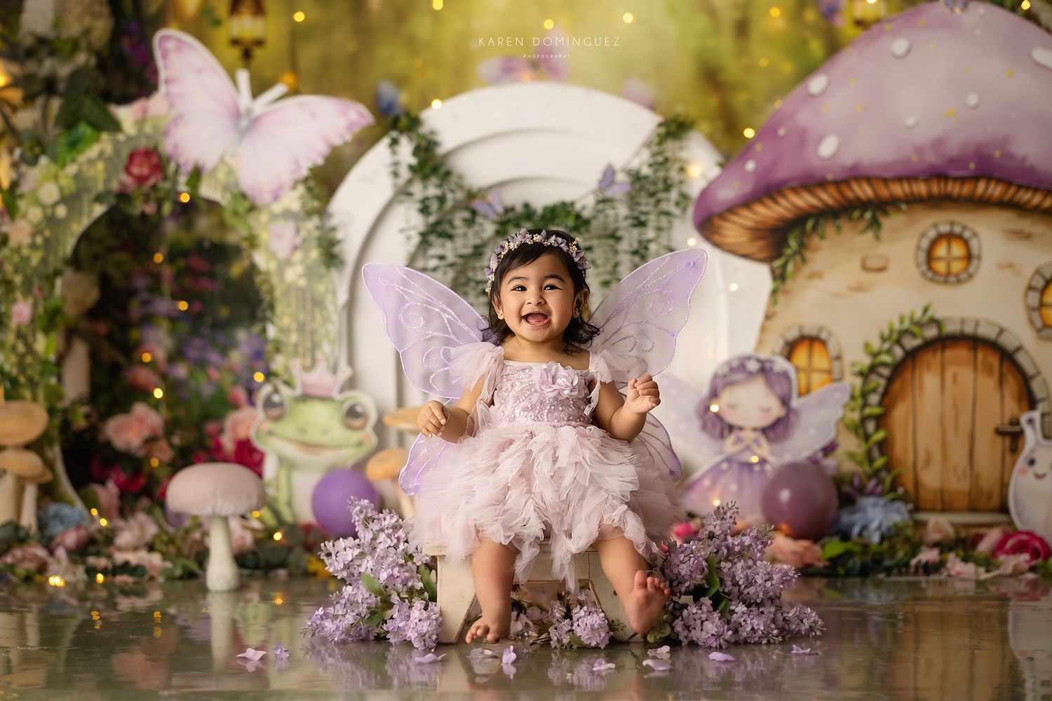 Toddler girl in fairy costume with wings poses in enchanted mushroom garden setting with butterflies and flowers.