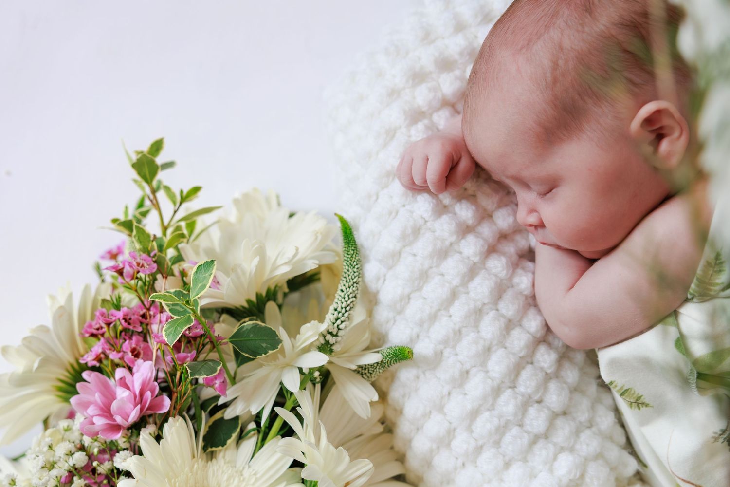 Sleeping newborn baby wrapped in white knit blanket beside a bouquet of white and pink flowers.