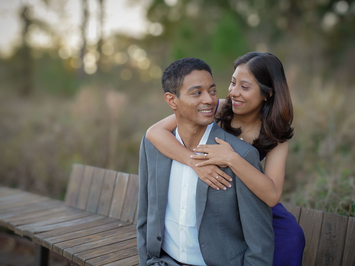 A couple embraces on a wooden bridge outdoors, the woman hugging the man from behind, both smiling warmly.