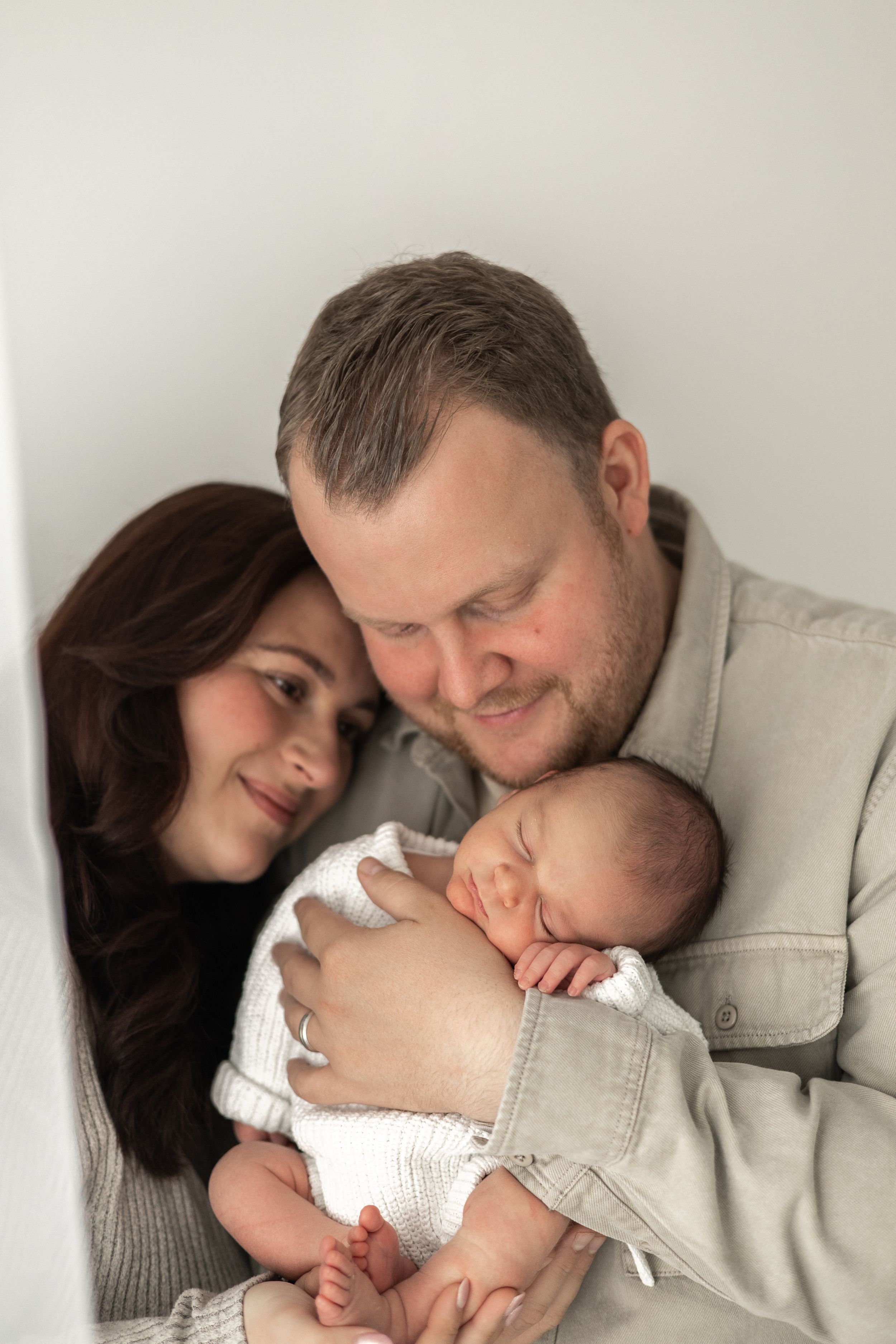 A couple smiles lovingly at their sleeping newborn baby wrapped in white, during Newborn baby photographer Nottingham studio