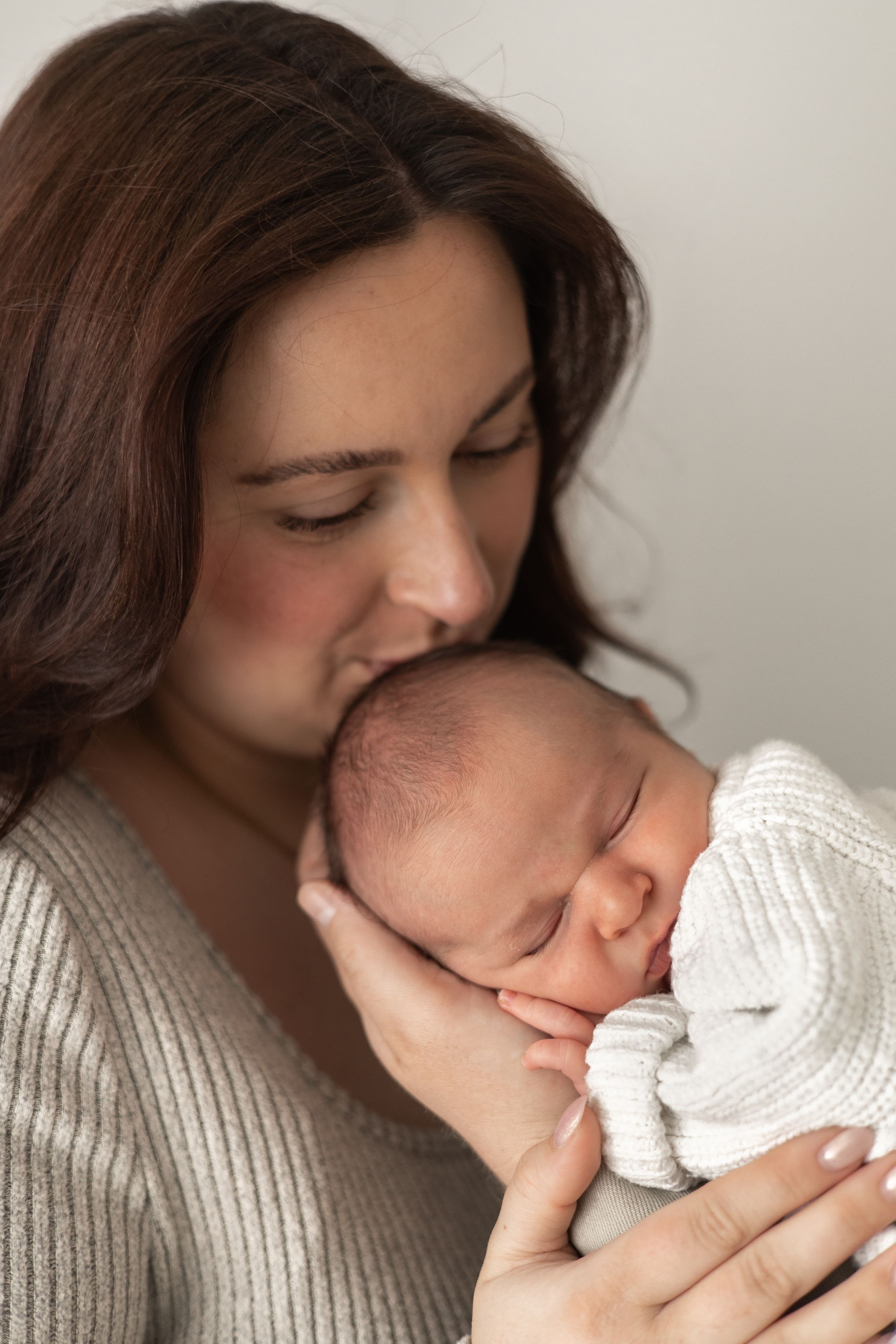 A tender mother gently kisses her sleeping newborn baby wrapped in a white knit blanket during Newborn baby photographer Nottingham studio
