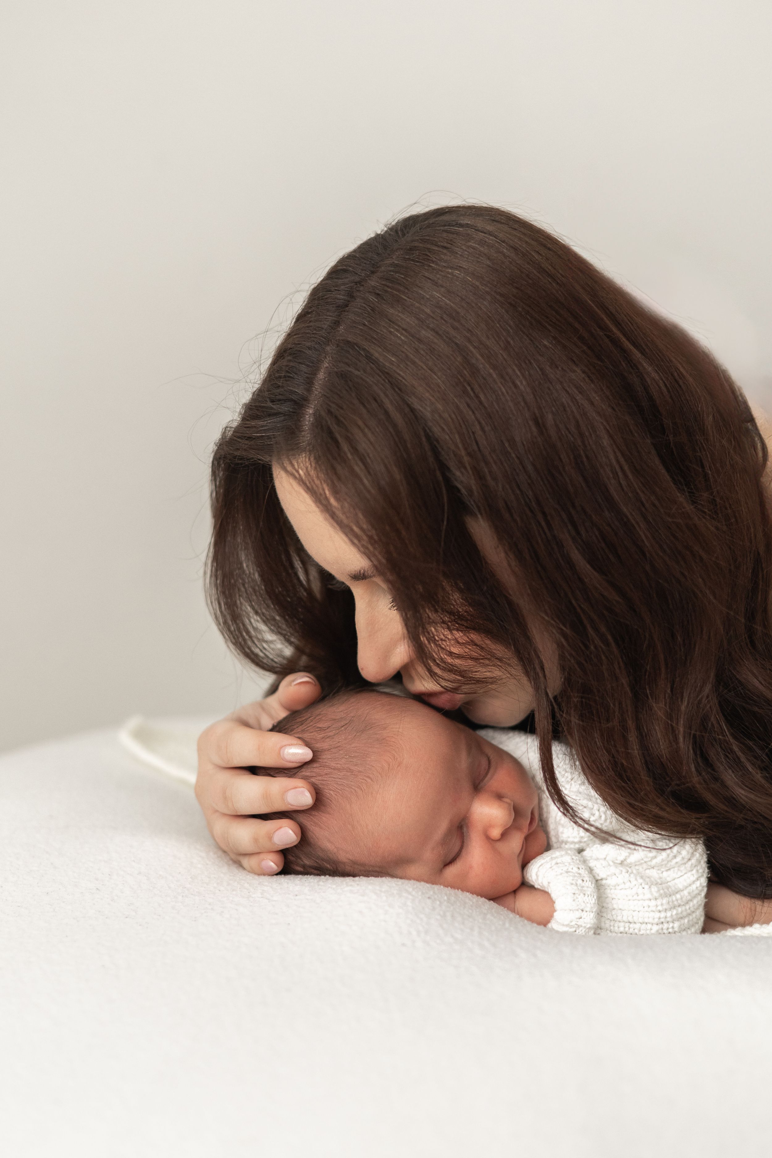 A mother with long dark hair tenderly kisses her sleeping newborn baby on a white surface during Newborn baby photographer Nottingham studio