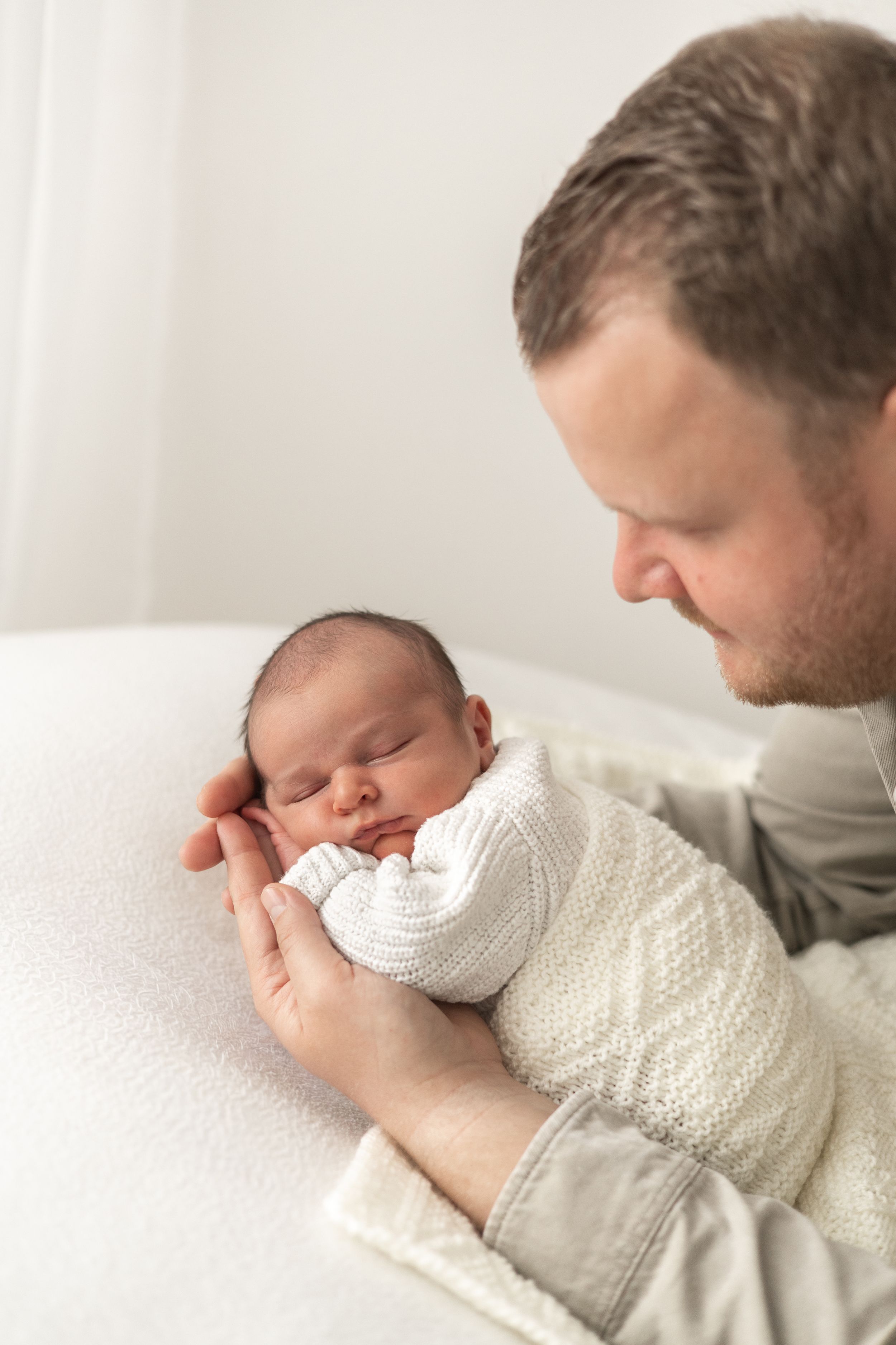 Father tenderly cradling sleeping newborn baby wrapped in white knit blanket during Newborn baby photographer Nottingham studio
