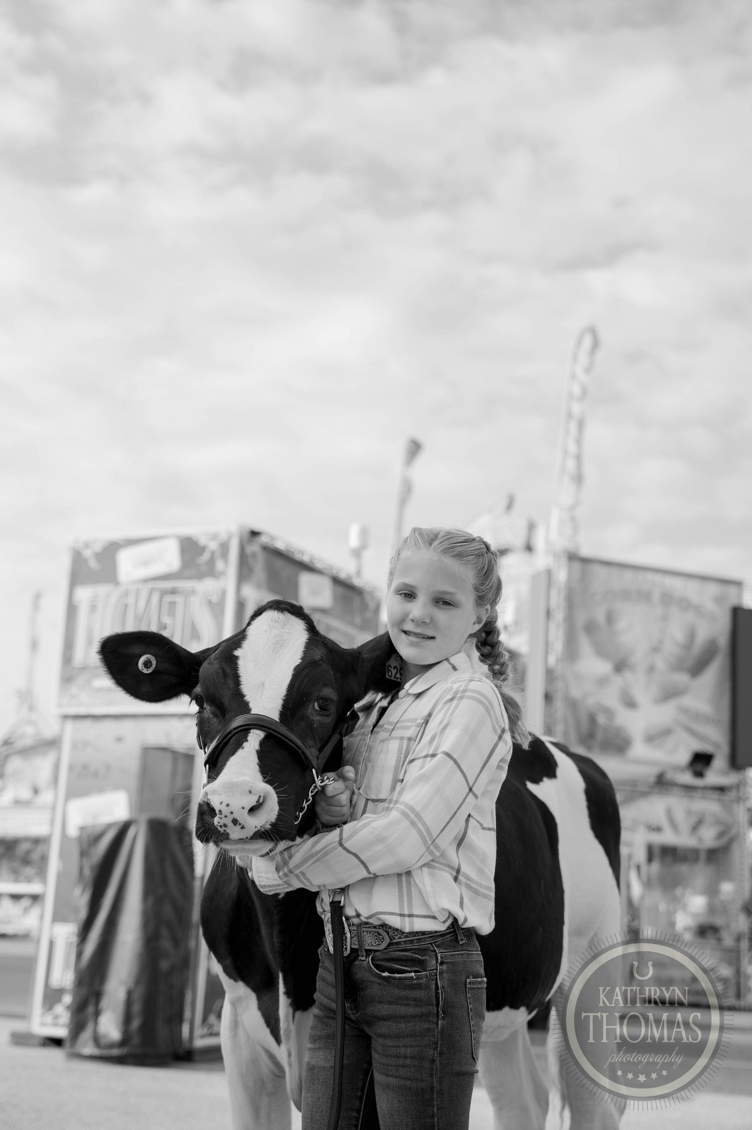 A young girl hugs a black and white dairy cow at a county fair with colorful carnival booths in the background.