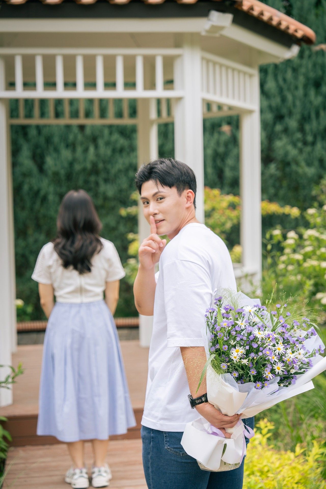 Young man hiding bouquet of purple flowers behind his back, gesturing silence, while woman stands near garden gazebo.
