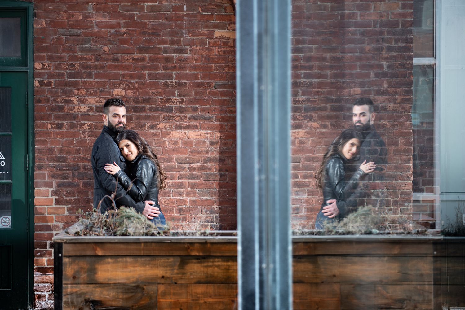 Couple hugging reflected in a window against a red brick wall in an urban setting.