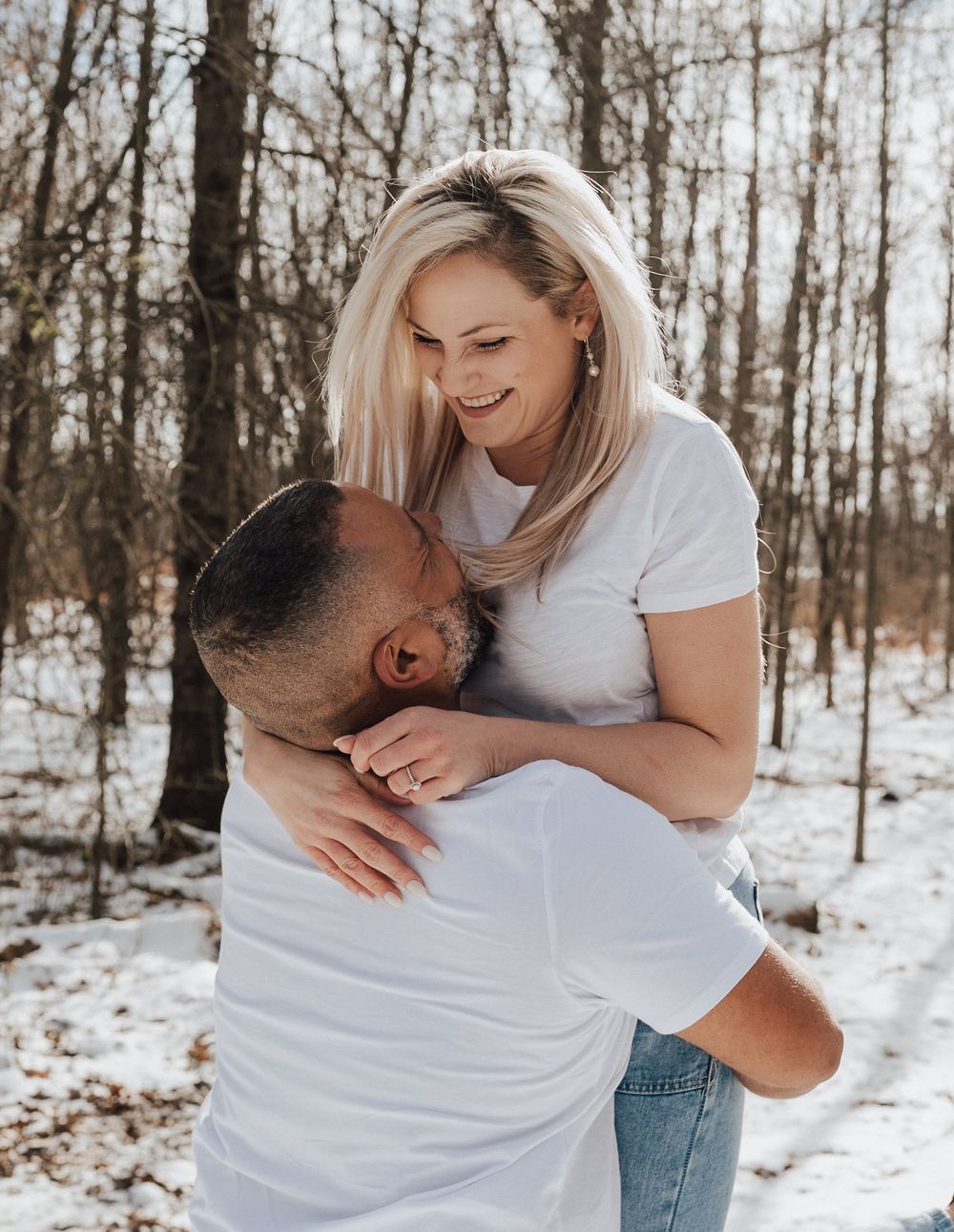 A couple shares a romantic embrace in a snowy winter forest, woman smiling as man hugs her in matching white shirts.