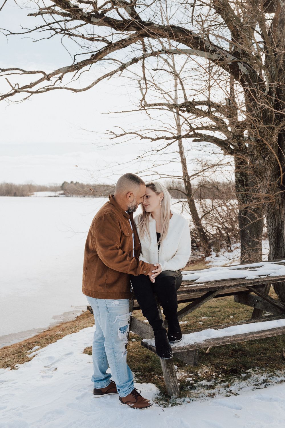 Couple sharing a romantic moment by a snowy lakeside, sitting on a weathered picnic table under bare winter trees.