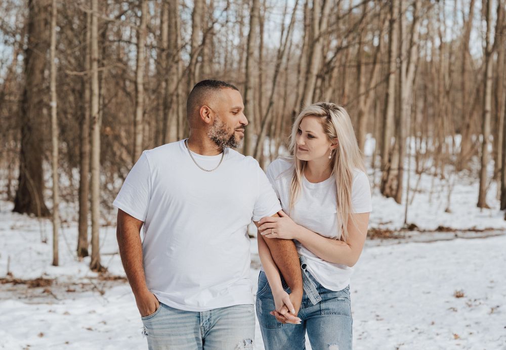 Couple in matching white t-shirts and jeans smiling at each other in a snowy winter forest setting.