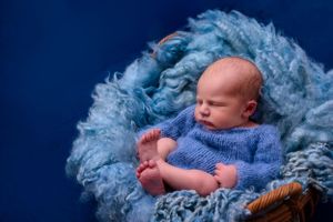 Infant in blue knit outfit resting on fluffy blue textile with dark blue backdrop.