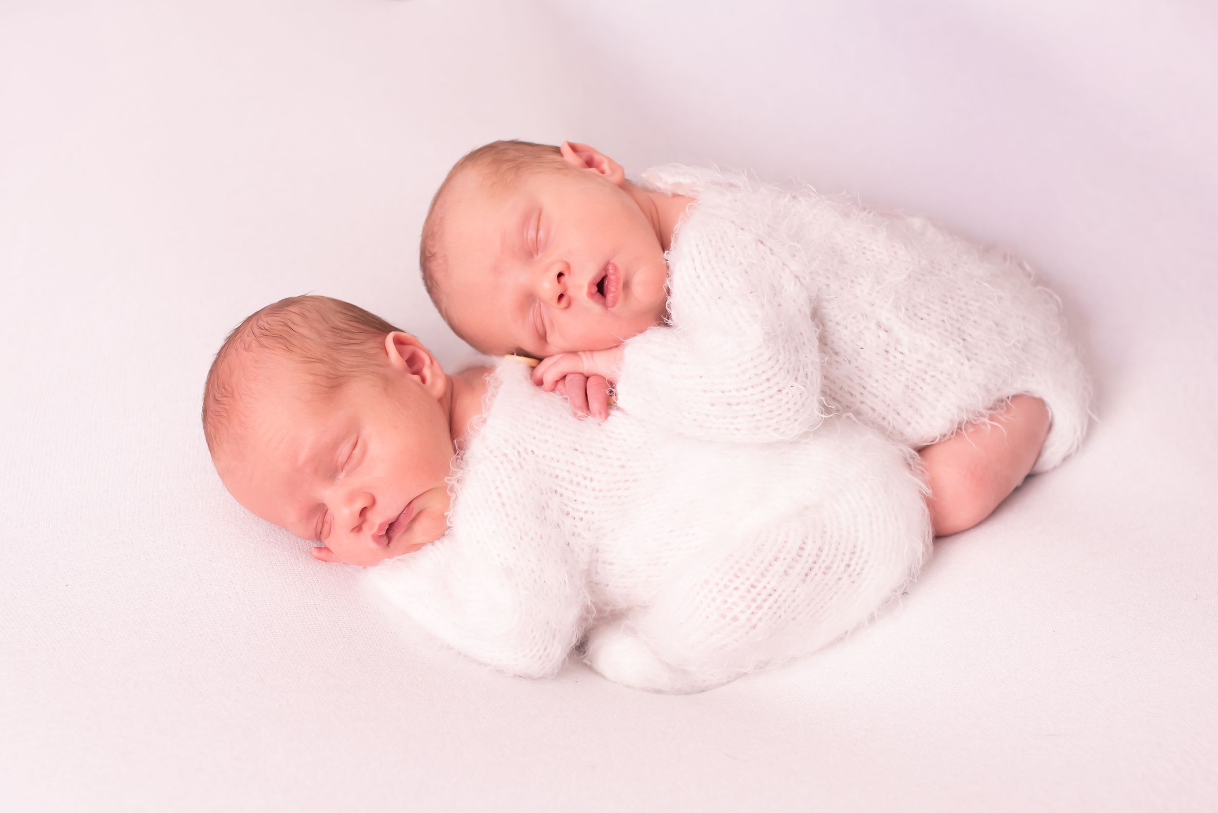 Two newborn twins sleeping peacefully together wrapped in white knit outfits on a soft white background.