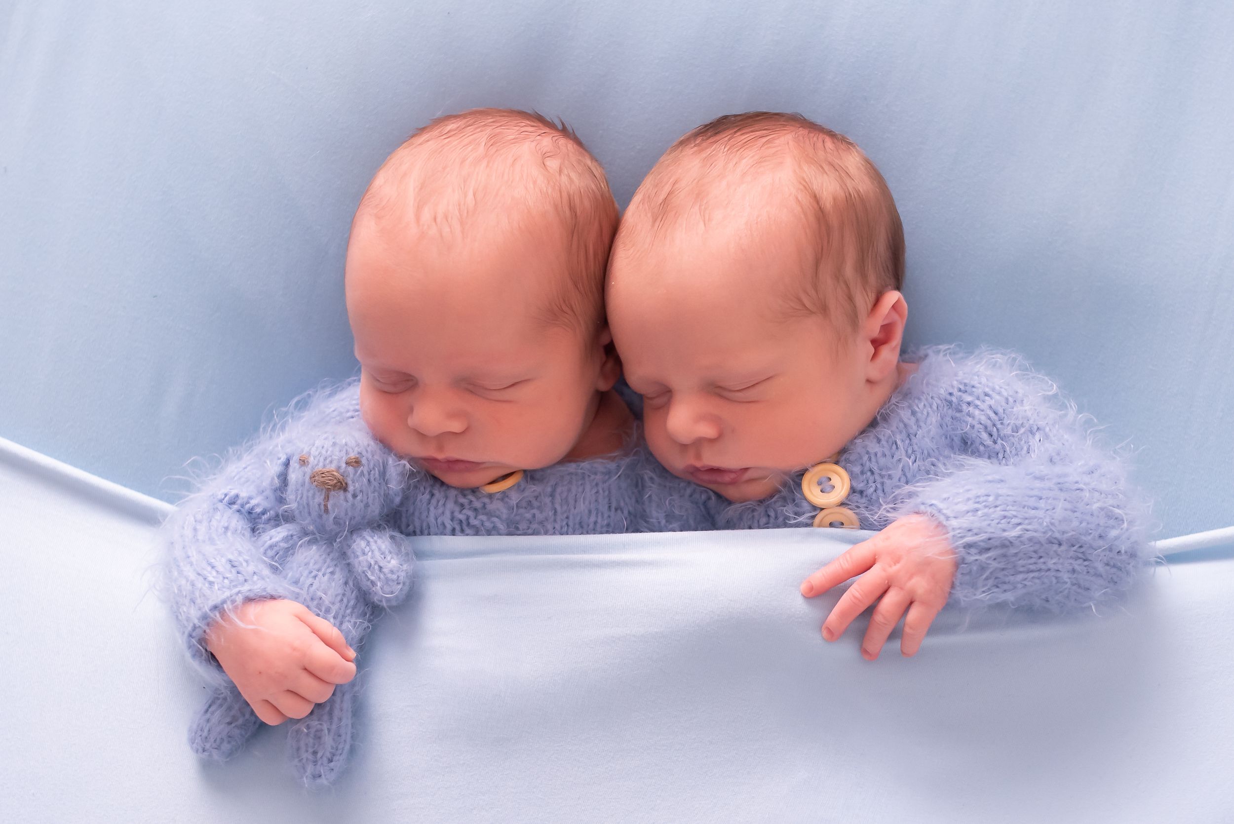Two sleeping newborn twins in matching blue fuzzy outfits rest side by side in a soft blue setting.