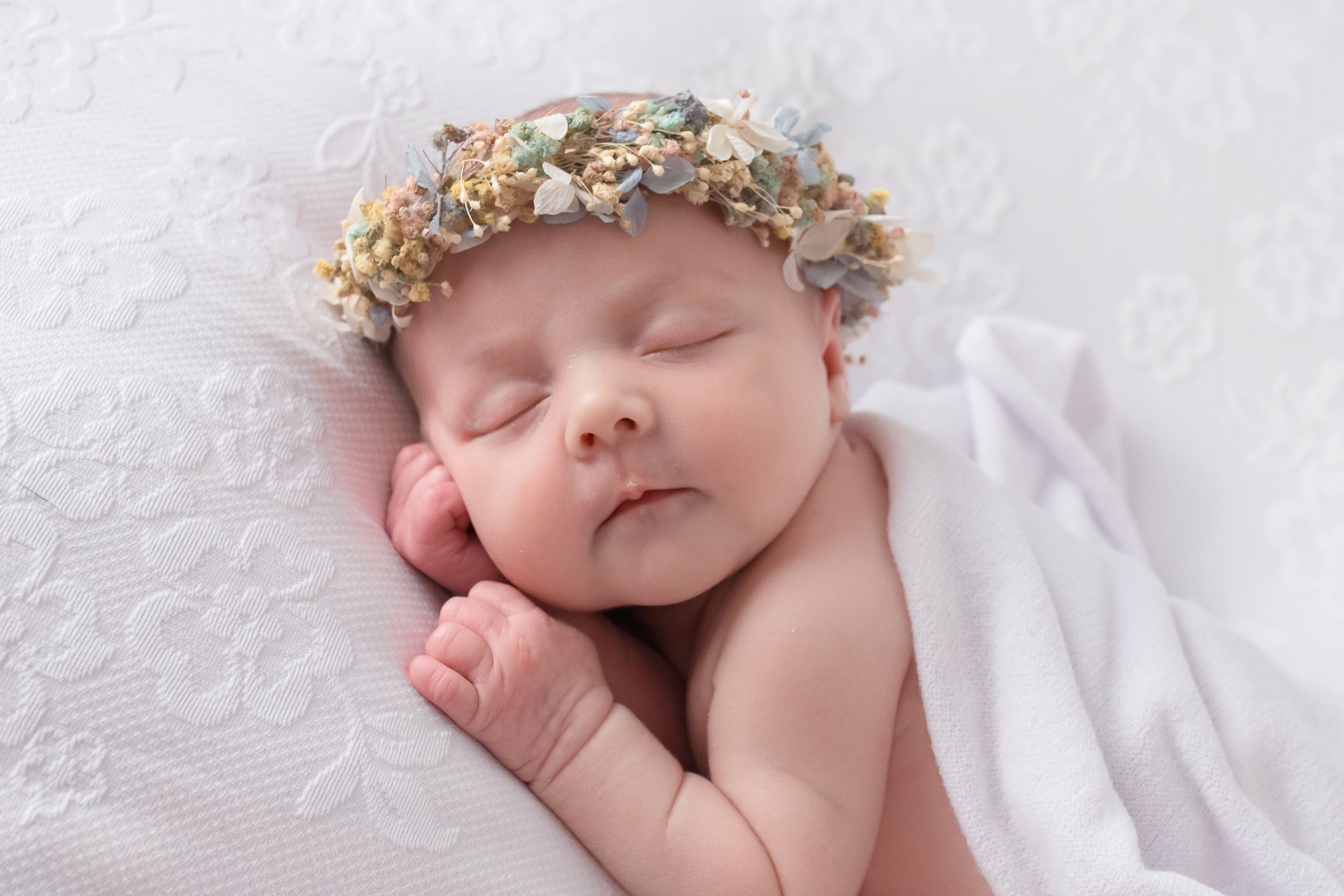 Peaceful newborn baby sleeping with a delicate floral crown, wrapped in a soft white blanket on a white textured background.