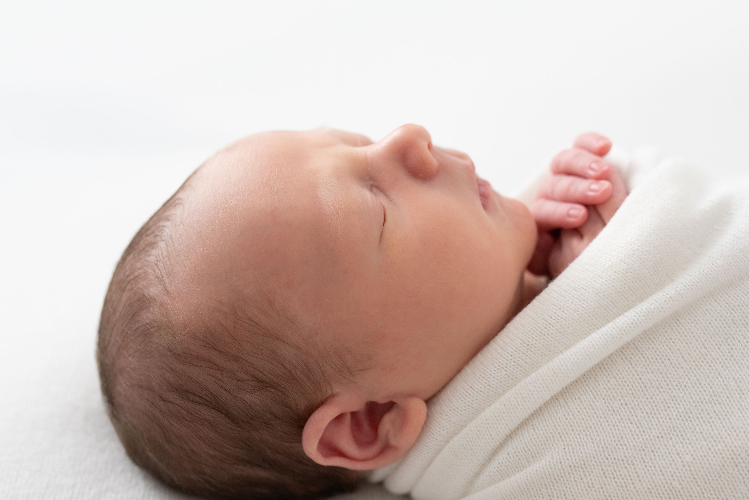 Peaceful newborn baby swaddled in white blanket sleeping, close-up profile view of infant with dark hair.