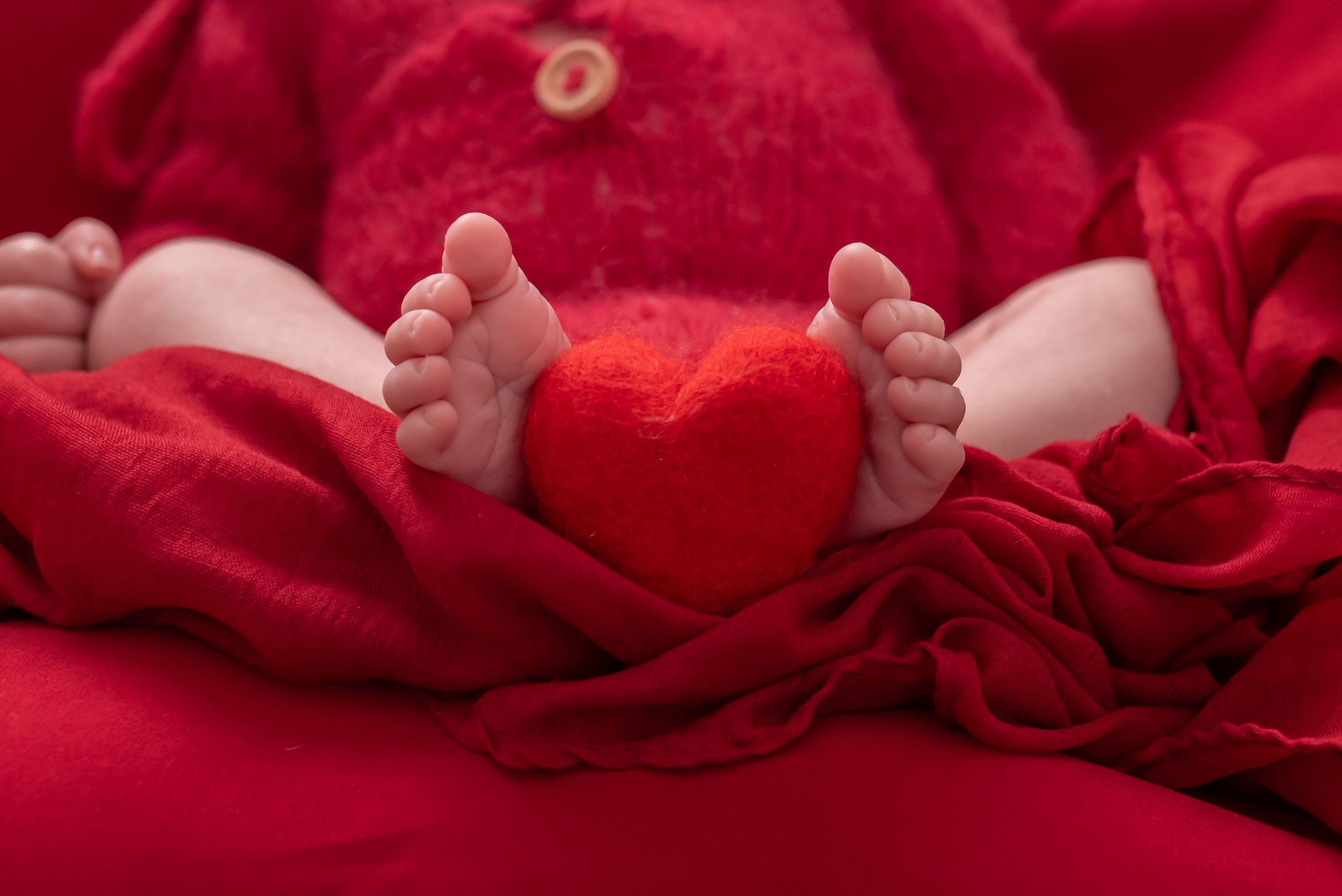 Close-up of newborn baby feet holding a red heart between them, wrapped in soft red blanket.