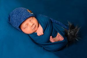 Swaddled newborn in blue wrap and knit cap sleeping on blue backdrop.