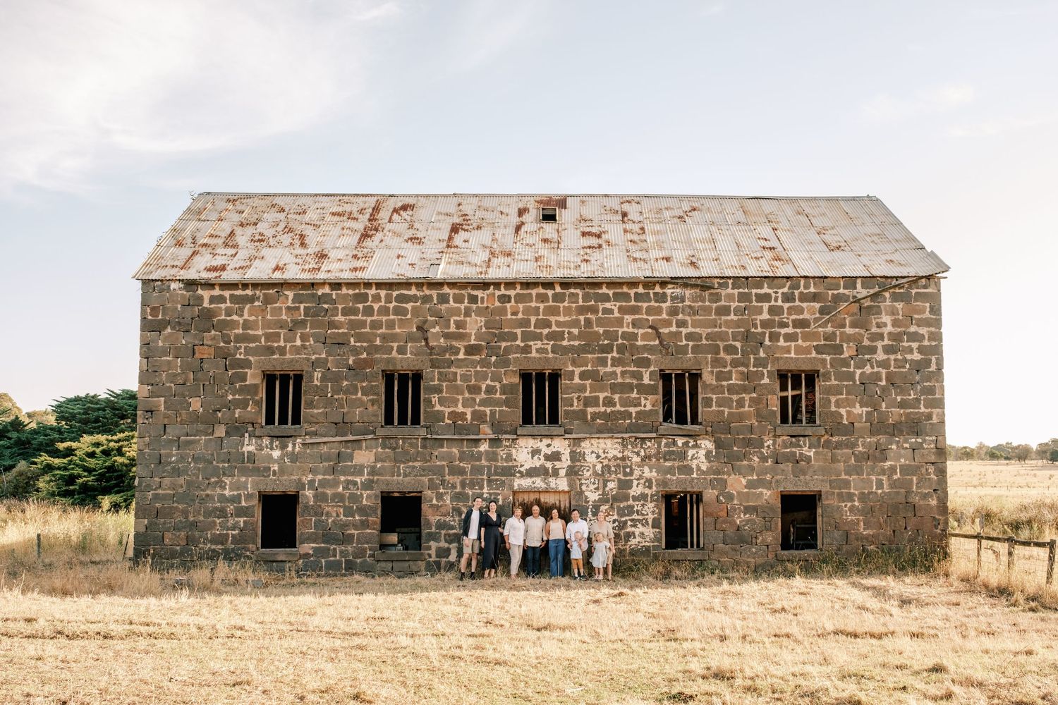 Group of people posing in front of a large historic stone barn building in a dry rural field.