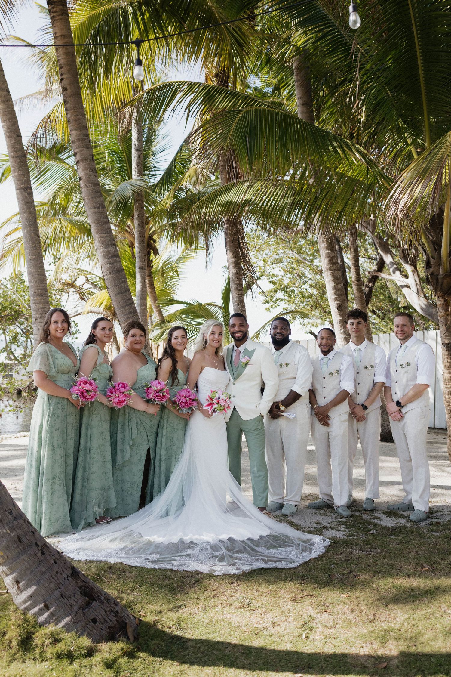 Wedding party poses under palm trees, bridesmaids in sage green gowns with pink bouquets, groomsmen in white suits.