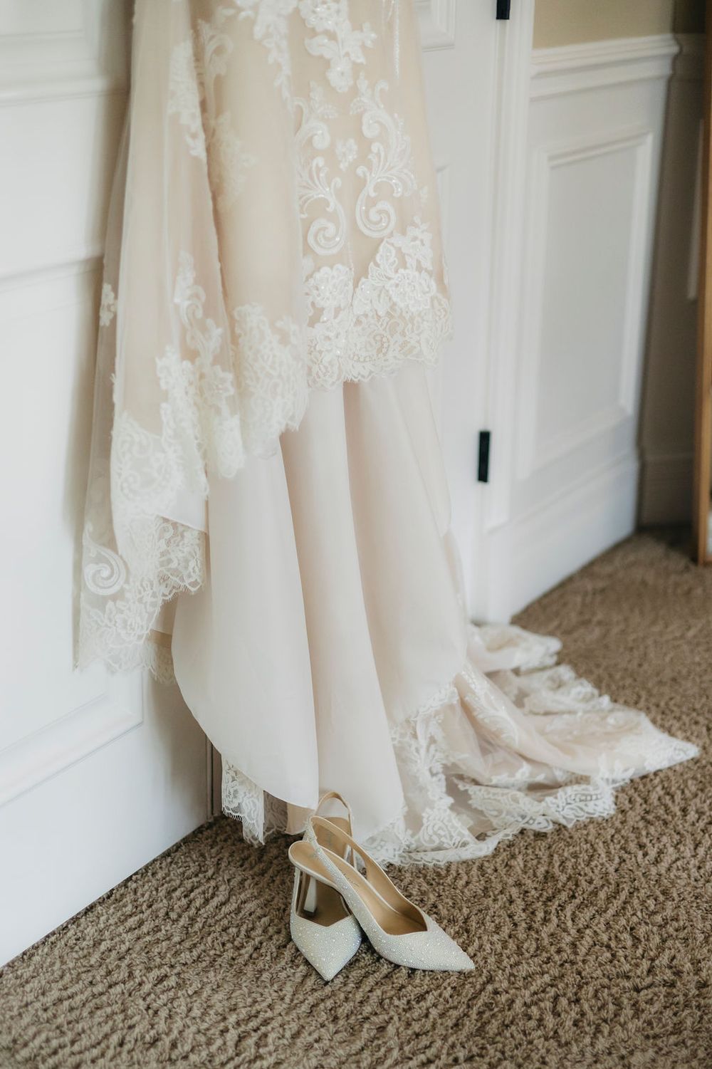 Elegant lace wedding dress hanging with white slingback heels on carpet below, bridal preparation detail shot in Chicago