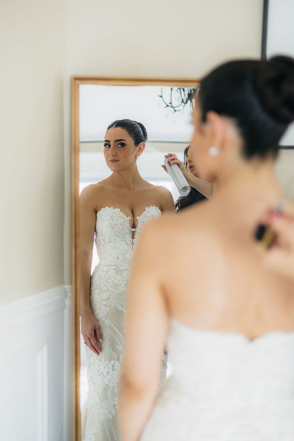Bride in lace strapless wedding gown getting ready, reflected in gold-framed mirror while stylist applies finishing touches in Chicago