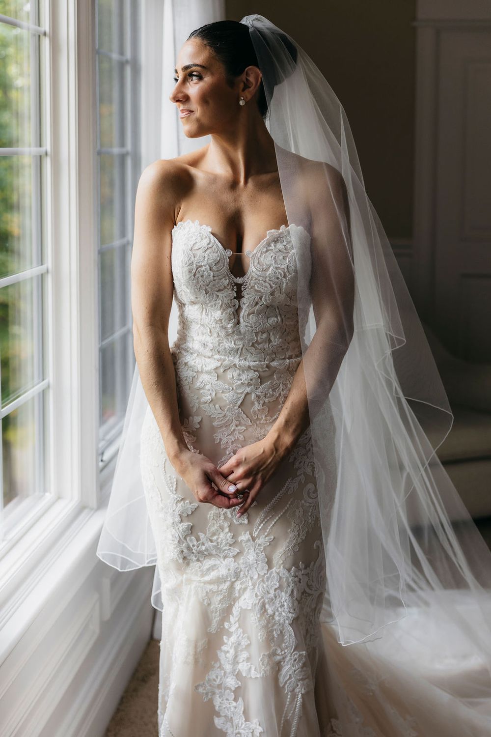 Bride in strapless lace wedding gown and flowing veil stands by window, gazing softly into the distance in Chicago while getting ready
