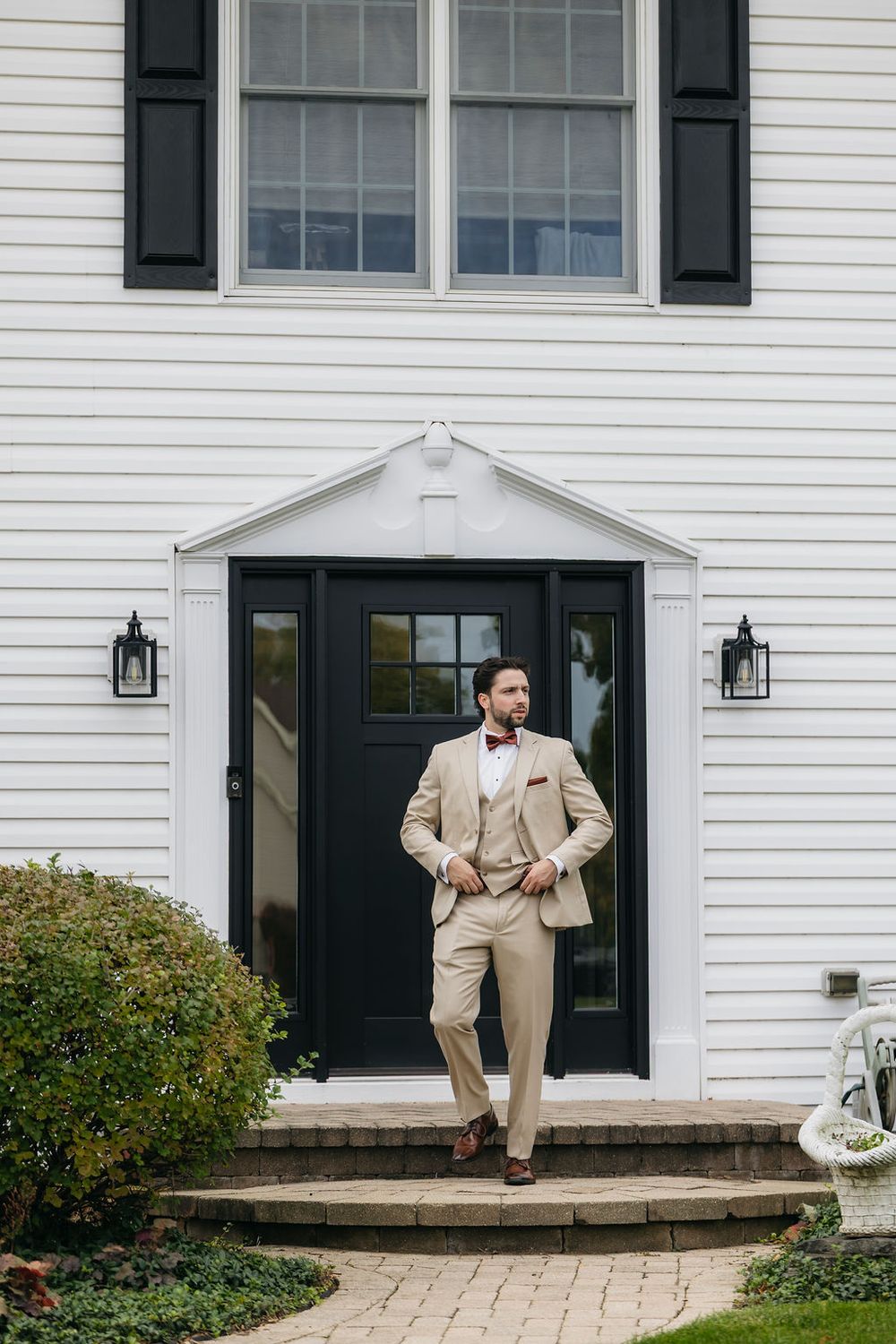 Elegant groom in tan suit with bow tie stands on steps of white colonial home entrance in Chicago