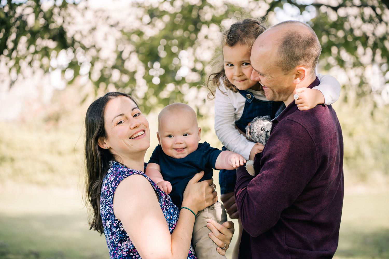 Happy family of four posing outdoors, parents holding two young children, smiling in a lush green park setting.