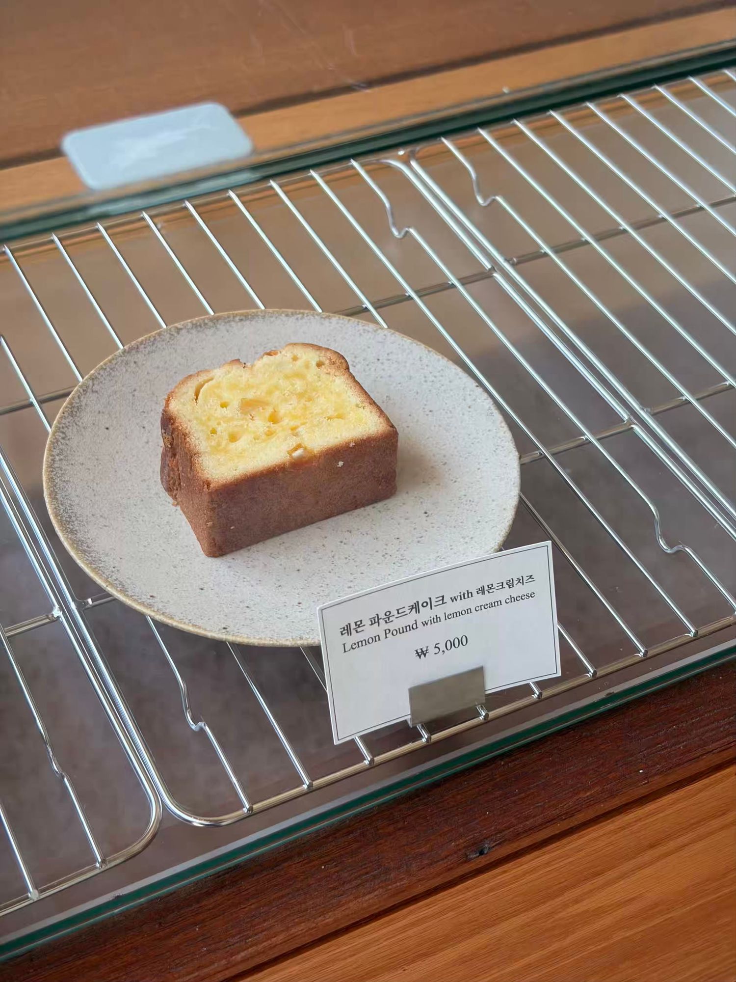 Slice of lemon pound cake with cream cheese on a ceramic plate displayed in a glass bakery case, priced at 5,000 won.