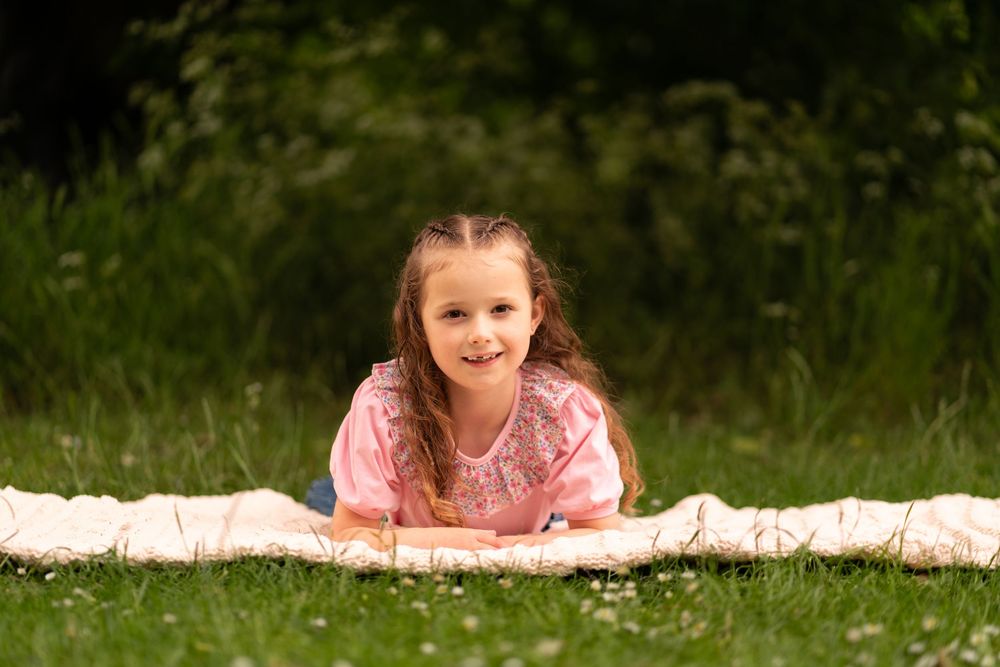 Young girl in pink top lying on blanket in grass, smiling at camera outdoors during a childhood photoshoot in Lincolnshire.